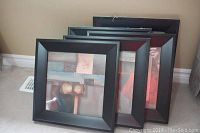 Set of 5 stacked framed prints with black frames resting against a wall.