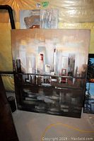 Photo showing two abstract embossed paintings on canvas leaning against a basement wall, one large 50x40 inches and a smaller 60x20 inches piece, depicting stylized cityscapes in earth tone colors.