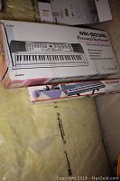 Box of Optimus MK-8038L personal keyboard and YKS2000B stand placed one atop another, showing keyboard box mostly front side with product illustration.