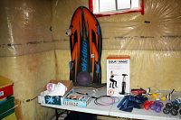 Wide view of the sports and exercise equipment on two white folding tables inside insulated room with yellow batting on walls. Visible items include Ski-Doo powder board, Squat Magic exerciser box, badminton rackets, hand weights, swim goggles, and other accessories.