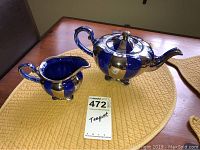 Blue and silver Arthurwood teapot and creamer set on a yellow placemat on a wooden table, with lot tag showing teapot labeling.