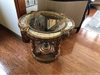 Side angled photo showing wood carvings, glass top, marble-like inset surface, and heavy build on hardwood floor next to a chair.