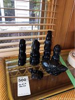Six black onyx sculptures displayed on a glass table with a bamboo-style frame and window blinds in background. Includes five tall totem-like sculptures and one leaf-shaped sculpture.