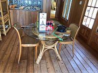 Round rattan table with beveled edge glass top and two matching rattan chairs with woven cane seats and backs in a wood paneled room.
