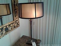 Lamp on marble table with ornate gold-framed mirror behind. Beige drum shade with black trim is lit showing fabric texture. Metal base with square footed bottom visible.