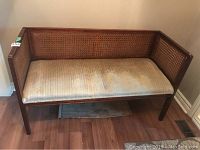 Front view of wood framed bench with cane sides and backrest, upholstered light tan seat cushion with wear marks