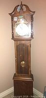 Front full view of the tall wooden Craftique Craftline grandfather clock against a wall, showing the clock face, decorative wooden top, and glass door with pendulum.