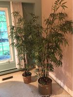 Photo showing two tall faux bamboo plants in different pots near a wall and window with vertical beige curtains, plants about 75 inches tall
