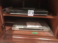 Both devices shown on a shelf: slim black REI DVD player on the top shelf, silver JVC VCR on lower shelf with manuals on top.