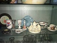 Photo of shelf with a child's ceramic tea set on tray including teapot, sugar bowl, creamer, and two teacups with saucers, Paragon teacup and saucer, commemorative 1939 Canada visit teacup and saucer, and other assorted small dishware and glass items.