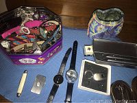 Photo of tin container filled with assorted wristwatches, costume jewellery, spectacles, wallets, and other accessories on a blue surface.
