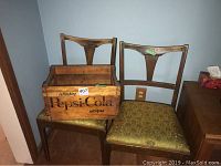 Photo of vintage Pepsi Cola wooden crate sitting on a wooden chair, alongside another similar wooden chair, against a blue wall.