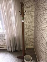 Tall wooden coat rack standing near a window with sheer curtains and sheet music patterned wallpaper. Shows detail of hooks and wood finish.