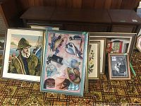 Overall view of multiple framed textile and art prints leaning against a cabinet, showing various images including a sailor man and pastel textile art with hats and birds.
