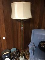 Vintage brass floor lamp approximately 64 inches tall with large cream fabric shade. Positioned beside a blue upholstered chair and wooden paneled wall.