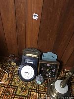 Lot of 7 clocks placed on patterned carpet with wood panel background, showing variety of shapes, materials, and sizes.