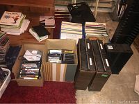 Overall view showing multiple boxes and crates containing vinyl albums, CDs, DVDs, and PlayStation games arranged on floor.