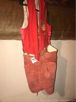 Photo of heavy suede lederhosen in reddish-brown color with front flap and suspenders paired with a red polka dot vest with emblem on chest hanging on a black hanger.