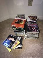 Stacks of approximately 40 music CDs in jewel cases on carpeted floor. Titles include Susan Boyle, The Canadian Tenors, The Kingston Trio visible on top of some stacks.