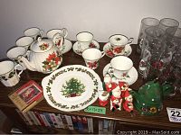 Christmas-themed dinnerware including teapot, cups, saucers, bowls with poinsettia and holly designs, a plate with Christmas tree and holly border, and several Santa and Christmas tree figurines arranged on a wooden shelf.