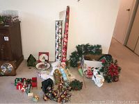 View of assorted Christmas decorations spread on floor including artificial greenery, figurines, wreaths, gift boxes, ribbons, and rolls of wrapping paper.