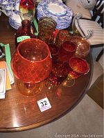 Thirteen assorted orange glassware pieces arranged on a wooden table including one large 9x3x12 inch stoppered vase, multiple goblets, tumblers and bowls.