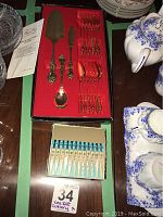 Photo of the gold plated Sealy 15-piece dessert set in a red lined box alongside the blue boxed 12-piece handles with floral decorated forks.