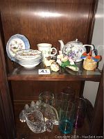 Photo of assorted bone china plates, teapot, ceramic figurines, and glassware on wooden shelf.