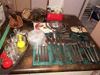 Overview of the lot showing the Paderno stainless steel cutlery laid out on a green cloth with knives, forks, spoons, and teaspoons, total of 20 pieces. Also visible are clear glass drinking glasses, red and yellow ornamental juice dispensers, a wooden duck decoration, metal trays, and a box containing China and glass mice collectibles.