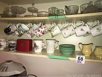 Shelf view of assorted bone china coffee mugs with floral prints, a red boxed Buckingham Palace mug, four green jadeite nappies, and clear glass serving dishes on upper shelf.