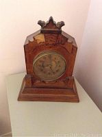 Front view of wooden antique clock showing carved top, round clock face with brass bezel, and damage at top front.