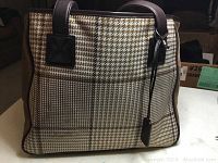 Side view of tote purse showing houndstooth and plaid pattern, leather handles, and tag.