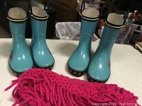 Two pairs of blue sunflower rain boots and pink sparkle fringe scarf arranged on a table.