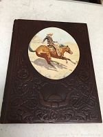Front cover of a brown leather bound collectible book titled 'The Cowboys', showing embossed floral patterns and a central oval color illustration of a cowboy on a horse.
