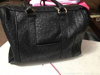 Front and side view of a large black embossed tote handbag with two short handles, showing the textured surface and front zipper compartment.