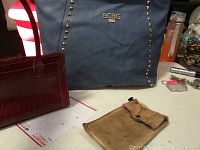 Overall view focusing on BCBG blue leather tote with stud detailing, Liz Claiborne ruby red mini handbag, and small beige cosmetic wallet on table.