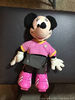 Front view of plush Minnie Mouse doll in pink shirt and grey skirt with pink skating shoes on black background.