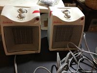 Two white Sunbeam portable electric heaters side by side showing front and top control dials with power cords