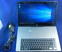 Front view of Acer Aspire R7 laptop open, powered on displaying Windows 10 Pro desktop with power cord beside it.