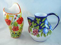 View of the front of vase with red and yellow floral hand paintings and the pitcher with blue, green, pink hand painted flowers and fruits