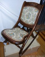 Front side view of vintage folding rocking chair showing floral upholstered seat and back rest, carved floral top detail, wood frame and curved rockers.