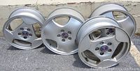 Four silver alloy Saab wheel rims displayed on ground, front view showing their three-spoke design and Saab logo cap.
