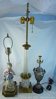 Three vintage lamps displayed against a white background. Tall brass lamp with glass midsection and two bulb sockets. Ceramic figurine lamp of a woman on left. Small ornate metal lamp on right.