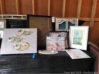 Eight nautical and coastal themed art pieces arranged on a black pallet including a fish painting, lenticular print, Wellfleet prints on wood, and sea paintings framed in varied styles.