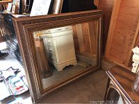 Large framed wall mirror resting against a wooden wall showing reflection of furniture and surroundings.