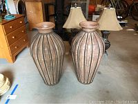 Photo of two large metal urns displayed on the floor indoors with other furniture and decor around.