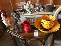 Entire lot displayed on wooden table includes ceramic rooster, sunflower bowls stacked on matching underplate, glass decanters, Kimberly Hodges red pitcher and hot plate, and assorted ceramic and glass items in background.