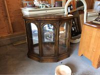 Front view of a wooden console cabinet with one decorative glass door and mirrored back panel, showing internal glass shelves with display items inside.