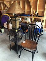 Photo showing the metal three-tiered end table with a large table lamp on top and next to a wooden side chair with black painted frame and natural wood seat.