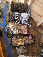 Box and trays containing long faux gemstone and pearl necklaces, mostly in shades of white, black, and multicolor beads, displayed on a wooden table in a garage setting.